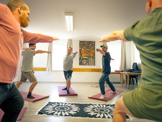 Ein Männer-Yogakurs in einem hellen Raum. Die Teilnehmer stehen auf bunten Matten und führen eine Armübung aus. Im Hintergrund ist ein farbenfrohes Gemälde zu sehen.