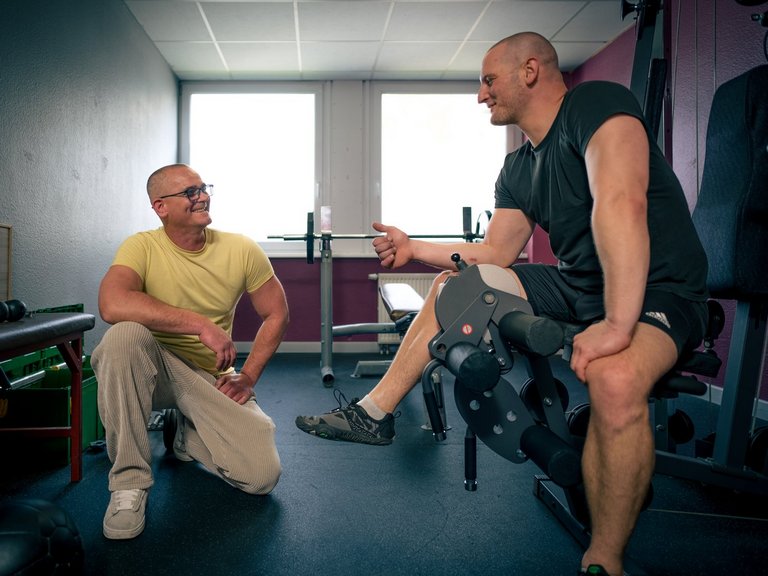 Zwei Männer unterhalten sich in einem Trainingsraum. Ein Mann im gelben T-Shirt kniet lächelnd auf dem Boden, während der andere Mann auf einem Trainingsgerät sitzt, ein Kniebandage trägt und gestikulierend spricht.