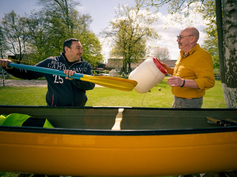 Zwei Männer stehen im Freien neben einem gelben Kanu. Der Mann links hält ein Paddel, während der Mann rechts Wasser aus einem weißen Eimer auf die Paddelklinge schüttet, vermutlich zum Reinigen oder Vorbereiten.