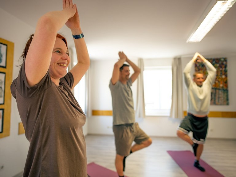 Drei Personen üben in einem hellen Raum Yoga. Eine lächelnde Frau im Vordergrund und zwei Männer im Hintergrund balancieren in der Baums pose auf einer Matte mit erhobenen Armen.