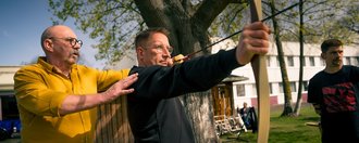 Ein Mann in schwarzem Oberteil spannt einen Bogen, während ein Mann im gelben Hemd ihn als Trainer anleitet und seine Haltung korrigiert.