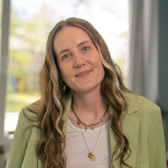 Janina-Sophie Roettger Porträt einer lächelnden Frau mit langen, welligen Haaren, die auf einem Schreibtisch in einem Büro sitzt. Sie trägt einen hellgrünen Anzug über einem weißen Top und mehrere Halsketten. Im Hintergrund sind ein Fenster und Büromöbel zu sehen.