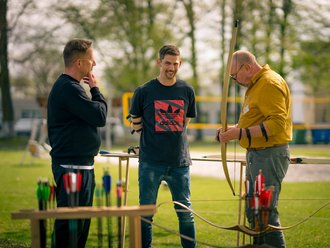 Drei Männer stehen auf einer Wiese beim Bogenschießen. Ein Mann im gelben Hemd hält einen Bogen, während ihn ein anderer Mann im schwarzen T-Shirt anlächelt. Ein dritter Mann beobachtet das Gespräch. Im Vordergrund stehen Pfeile in Haltern.