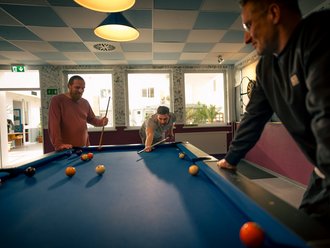 Drei Männer spielen eine Partie Poolbillard. Ein Mann beugt sich über den blauen Tisch, um einen Stoß auszuführen, während die anderen beiden ihm aufmerksam zuschauen.