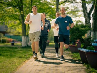 Vier Männer in Sportkleidung joggen gemeinsam auf einem gepflasterten Weg durch einen grünen Park an einem sonnigen Tag.