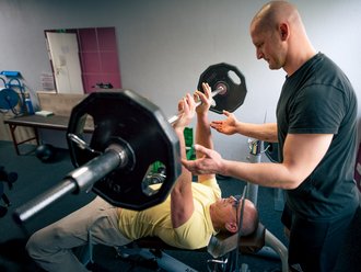 Ein Mann liegt auf einer Trainingsbank und drückt eine Langhantel in die Höhe. Ein Trainer steht daneben, überwacht die Bewegung und sichert ihn mit den Händen.