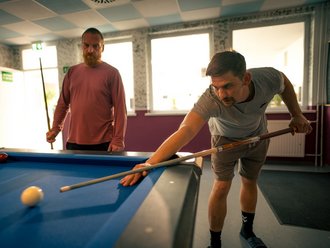 Zwei Männer spielen Billard. Ein Mann im grauen T-Shirt beugt sich konzentriert über den blauen Tisch und zielt mit dem Queue. Im Hintergrund steht ein bärtiger Mann und beobachtet das Spiel.