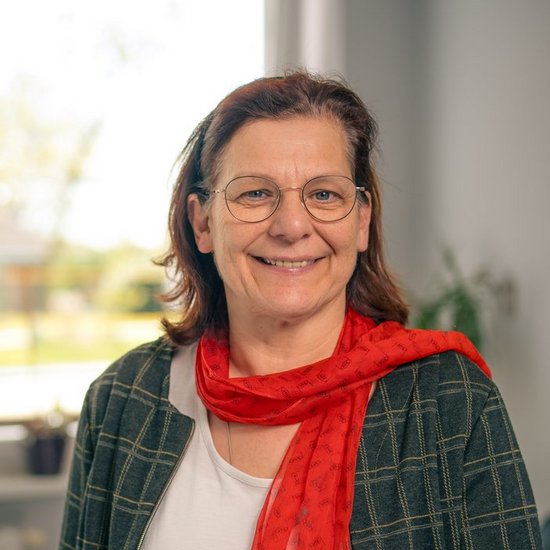 Eine lächelnde Frau mit Brille und rotem Schal steht in einem Büro. Sie trägt eine dunkelgrüne karierte Jacke über einem weißen Oberteil. Im Hintergrund sind unscharf ein Fenster und Büromöbel zu erkennen.