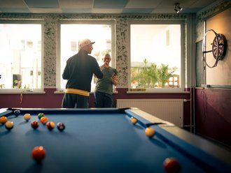In einem Spielzimmer steht im Vordergrund ein Billardtisch mit Bällen. Im Hintergrund unterhalten sich zwei Männer vor Fenstern, während links eine Frau Darts hält. Rechts an der Wand hängt ein Dartbrett.