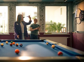 Im Vordergrund liegt ein blauer Billardtisch mit bunten Kugeln. Dahinter werfen drei Personen Dartpfeile. Eine Frau in einem lila Blazer wirft gerade einen Pfeil, während ein Mann daneben zusieht. Links steht eine weitere Frau. Der Raum hat große Fenster.