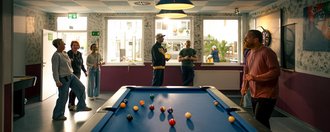 Ein blauer Billardtisch steht im Vordergrund. Ein Mann mit Queue bereitet einen Stoß vor, während eine Gruppe junger Menschen im Hintergrund steht, zuschaut und miteinander spricht.