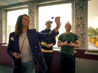 Eine Frau in einem lila Blazer hält einen Dartpfeil hoch und zielt. Im Hintergrund stehen zwei Männer, die das Spiel beobachten; einer hält weitere Darts bereit, während der andere lächelnd ein Smartphone in der Hand hält.