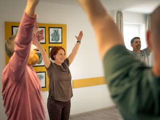 Eine lächelnde Frau mit roten Haaren steht im Mittelpunkt einer Gruppe und hebt beide Arme nach oben. Links und rechts von ihr sind weitere Personen zu sehen, die die gleiche Bewegung ausführen, vermutlich bei einer Yoga- oder Gymnastikstunde in einem hellen Raum.