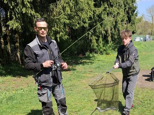 Zwei Männer stehen auf einer grünen Wiese vor hohen Nadelbäumen. Der Mann links hält eine Angelrute, während der Mann rechts ein Kescher mit einem Fisch hält. Beide tragen funktionelle Outdoor-Kleidung.