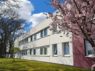 Ein weißes, zweistöckiges Gebäude mit der Aufschrift „The Park Hotel OPEN“. Im Vordergrund blüht ein großer Kirschbaum auf einer grünen Wiese unter einem blauen Himmel mit Wolken.