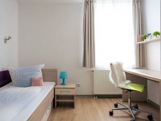 Ein helles Einzelzimmer mit hellem Holzboden. Links steht ein Bett mit blauer Bettwäsche und ein Nachttisch mit einer blauen Lampe. Rechts befindet sich ein Schreibtisch unter einem Regal mit kleinen Pflanzen. Davor steht ein grüner Bürostuhl mit weißem Fellüberwurf. Ein großes Fenster mit Vorhängen sorgt für viel Tageslicht.