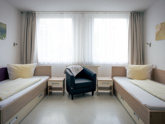 Ein helles Schlafzimmer mit zwei Einzelbetten, die mit gelber Bettwäsche bezogen sind. In der Mitte steht ein dunkelblauer Sessel zwischen zwei Nachttischen. Im Hintergrund befindet sich ein großes Fenster mit weißen Vorhängen.