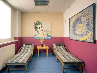 Ein Ruheraum mit zwei Holzliegestühlen auf blauem Fliesenboden. Dazwischen steht ein kleiner Holztisch mit einer Pflanze. An den Wänden hängen ein Pop-Art-Gemälde einer Frau und ein großes abstraktes Mosaikbild.