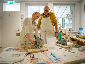 In einem hellen Atelier arbeiten ein Mann und eine Frau gemeinsam an einer Skulptur aus Papiermaché. Beide tragen schmutzige weiße Schürzen. Die Frau formt das Objekt, während der Mann sie beobachtet. Der Tisch ist mit Farben und Werkzeug bedeckt.