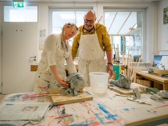 In einem hellen Atelier arbeiten ein Mann und eine Frau gemeinsam an einer Skulptur aus Papiermaché. Beide tragen schmutzige weiße Schürzen. Die Frau formt das Objekt, während der Mann sie beobachtet. Der Tisch ist mit Farben und Werkzeug bedeckt.