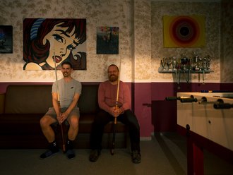 Zwei Männer sitzen lächelnd auf einem braunen Sofa und halten Billard-Keulen. Hinter ihnen an der Wand hängt ein großes Pop-Art-Porträt einer Frau. Rechts im Bild befinden sich ein Regal mit Trophäen und ein Tischkicker.
