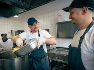 In einer Großküche füllt ein Koch Kartoffelwürfel aus einer Metallschale in einen großen Topf. Ein Kollege rechts im Bild beobachtet ihn lächelnd. Im Hintergrund ist eine weitere Mitarbeiterin in Kochkleidung zu sehen.