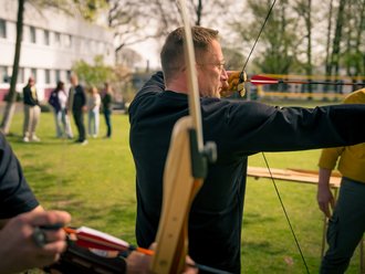 Ein Mann in einem schwarzen Pullover und mit Brille spannt einen Holzbogen und zielt mit einem Pfeil. Im unscharfen Hintergrund befinden sich weitere Personen auf einer grünen Wiese.