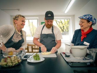 Drei Personen stehen in einer Küche und bereiten gemeinsam Obst zu. Ein Mann mit Schürze schneidet eine Banane, während eine Frau links und eine ältere Dame rechts lächelnd dabei zuschauen. Auf der Arbeitsplatte stehen Schüsseln mit geschnittenem Obst und Trauben.