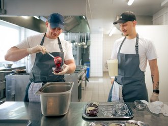 Zwei Köche in einer professionellen Küche bereiten Desserts zu. Einer füllt rote Fruchtsoße in Gläser, während der andere eine Kanne mit Sahne hält und lächelnd zuschaut.