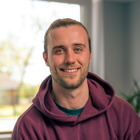 Baldur Philipp Ein junger Mann mit kurzem Haar und Bartstoppeln sitzt lächelnd in einem hellen Büro. Er trägt einen burgunderroten Hoodie und blickt freundlich direkt in die Kamera. Im Hintergrund sind ein Fenster und ein Schreibtisch unscharf zu erkennen.