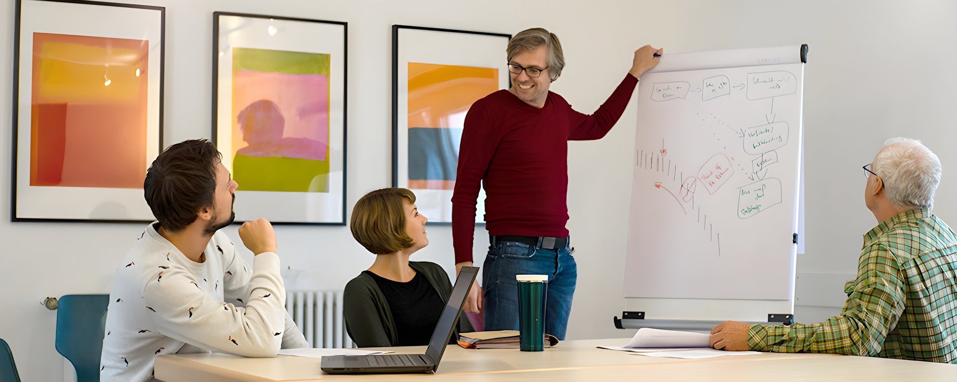 Besprechungsraum: ein junger Mann steht an einem Flipchart und erklärt einer kleinen Gruppe lächelnd sein "Gekritzel"
