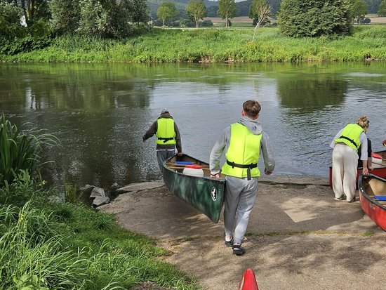 Drei Personen in leuchtend gelben Rettungswesten tragen Kanus zu einem Fluss. Zwei tragen gemeinsam ein grünes Kanu, während eine dritte Person ein rotes Kanu am Ufer vorbereitet. Im Vordergrund ist die Spitze eines weiteren roten Kanus zu sehen. Der Hintergrund zeigt grüne Ufer und bewaldete Hügel.