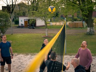 Fünf Männer spielen Volleyball auf einem Sandplatz im Freien. Ein gelb-blauer Ball schwebt über dem Netz, während ein Spieler ihn schlägt. Die anderen Männer beobachten das Spiel aufmerksam.
