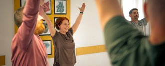 Eine lächelnde Frau mit roten Haaren steht im Mittelpunkt einer Gruppe und hebt beide Arme nach oben. Links und rechts von ihr sind weitere Personen zu sehen, die die gleiche Bewegung ausführen, vermutlich bei einer Yoga- oder Gymnastikstunde in einem hellen Raum.