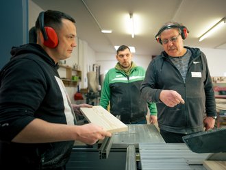Drei Männer in einer Holzwerkstatt. Ein Ausbilder mit Gehörschutz erklärt einem jungen Mann die Handhabung einer Kreissäge, während dieser ein Holzbrett positioniert. Ein weiterer Mann beobachtet die Situation im Hintergrund.