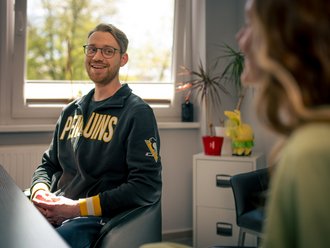 Ein junger Mann mit Brille und Bart sitzt lächelnd in einem Büro. Er trägt einen schwarzen Kapuzenpullover mit der gelben Aufschrift „Penguins“. Er blickt freundlich auf eine Frau im Vordergrund, die unscharf zu sehen ist, was auf ein Gespräch hindeutet.