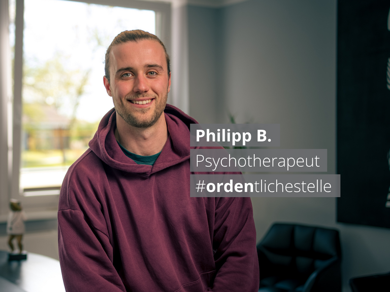 Porträt von Philipp B., einem lächelnden Psychotherapeuten in einem lila Hoodie. Ein Textoverlay nennt seinen Namen, Beruf und den Hashtag #ordentlichestelle vor dem Hintergrund eines hellen Büros.