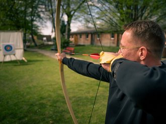 Ein Mann schießt mit einem Bogen auf eine Zielscheibe in der Ferne. Er trägt eine Brille und einen gelben Handschuh an der Zughand, während er den Pfeil auf einer grünen Wiese spannt.