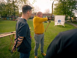 Ein Mann in einem gelben Hemd spannt einen Bogen und zielt auf eine Zielscheibe. Zwei weitere Männer beobachten ihn dabei auf einer grünen Wiese im Freien.