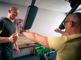 Ein Fitnesstrainer im schwarzen T-Shirt steht lächelnd neben einem Mann im gelben Shirt. Der Mann trainiert auf einer Maschine, während der Trainer ihm motivierend den Daumen hoch zeigt.