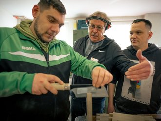 Drei Männer in einer Werkstatt. Ein junger Mann in grüner Jacke sägt Holz, während ein älterer Mann mit Gehörschutz ihn anleitet. Ein dritter Mann beobachtet aufmerksam. Die Szene zeigt eine praktische Unterweisung oder Ausbildung im Handwerk.