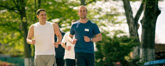 Zwei Männer joggen auf einem gepflasterten Weg in einem Park, gefolgt von zwei weiteren Läufern. Der Mann links trägt ein weißes ärmelloses Shirt, der Mann rechts ein blaues T-Shirt. Sie laufen an grünen Bäumen und Bänken vorbei.