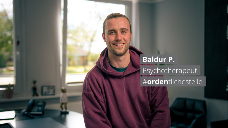 Ein lächelnder junger Mann in einem burgunderroten Hoodie sitzt vor einem Fenster. Ein Textfeld neben ihm zeigt: „Baldur P. Psychotherapeut #ordentlichestelle".