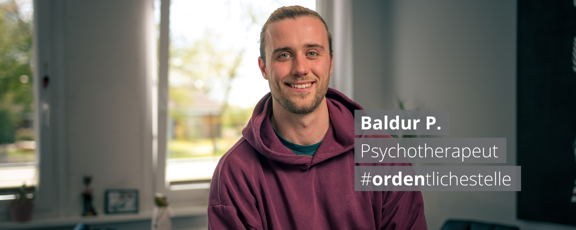 Ein lächelnder junger Mann in einem burgunderroten Hoodie sitzt vor einem Fenster. Ein Textfeld neben ihm zeigt: „Baldur P. Psychotherapeut #ordentlichestelle".