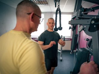 Ein Fitness-Trainer erklärt einem Kunden die Handhabung einer Trainingsmaschine. Der Trainer im schwarzen Shirt steht dem Kunden im gelben Shirt gegenüber und gestikuliert, um die richtige Übungsausführung zu demonstrieren.