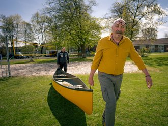 Ein kahlköpfiger Mann mit Schnurrbart und Brille auf der Stirn trägt gemeinsam mit einem zweiten Mann ein gelbes Kanu über eine Wiese.