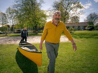 Ein kahlköpfiger Mann mit Schnurrbart und Brille auf der Stirn trägt gemeinsam mit einem zweiten Mann ein gelbes Kanu über eine Wiese.